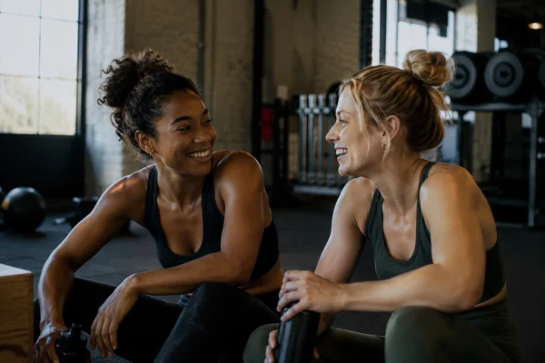 Två kvinnor skrattar tillsammans på gymmet efter ett träningspass – ett exempel på varför man ska träna med en vän