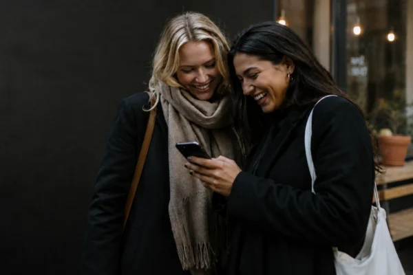 Två kvinnor skrattar tillsammans och tittar på en mobiltelefon – illustrerar hur enkelt det är att hitta vänner via vänskapsapp