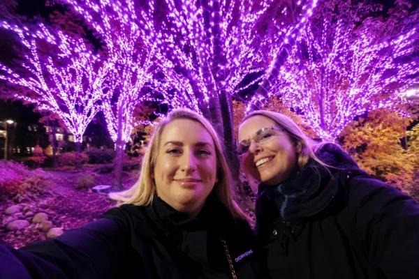 Två kvinnor i Göteborg tar en selfie tillsammans under upplysta träd efter att ha träffat nya vänner och byggt gemenskap.