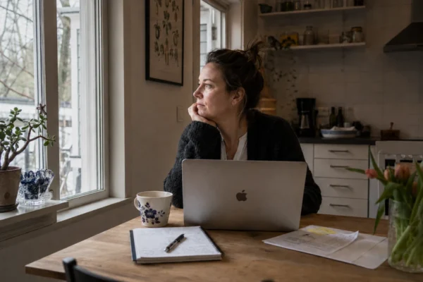 Kvinna sitter ensam med laptop vid köksbord och tittar ut genom fönstret – illustrerar ensamheten av att jobba hemifrån